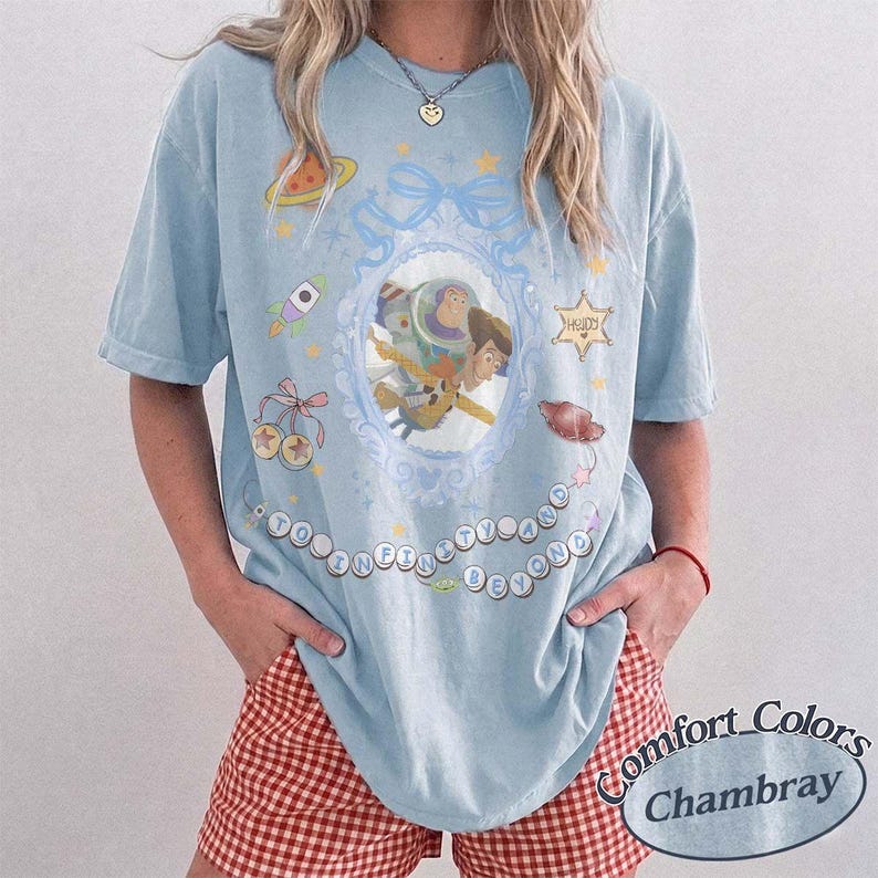 Peut inclure: Un t-shirt bleu clair avec un motif central repr&eacute;sentant Buzz l'&Eacute;clair et Woody dans un cadre ovale orn&eacute;. Le design comprend une fus&eacute;e, une plan&egrave;te, des &eacute;toiles et le texte TO INFINITY AND BEYOND &eacute;crit en cercle autour des personnages.