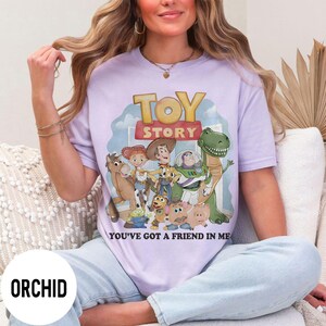 K&ouml;nnte beinhalten: Orchideenfarbenes T-Shirt mit einer Toy Story-Grafik. Das Design zeigt Woody, Buzz Lightyear und andere Charaktere aus dem Film, mit dem Text "TOY STORY" und "YOU'VE GOT A FRIEND IN ME".