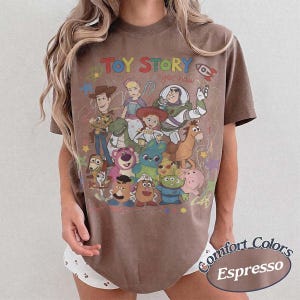 May include: A brown short-sleeve t-shirt featuring a graphic print of Toy Story characters including Woody, Buzz Lightyear, Jessie, and others. The text Toy Story is printed in colorful letters at the top, with the phrase yee haw written below.