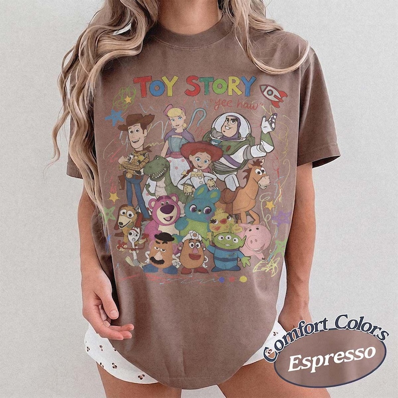 May include: A brown short-sleeve t-shirt featuring a graphic print of Toy Story characters including Woody, Buzz Lightyear, Jessie, and others. The text Toy Story is printed in colorful letters at the top, with the phrase yee haw written below.
