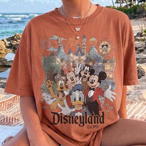 May include: A rust-colored short-sleeve t-shirt featuring a graphic print of Mickey Mouse, Minnie Mouse, Donald Duck, Goofy, and Pluto in front of a castle. The text "Disneyland Est. 1955" is printed in black at the bottom of the graphic design.