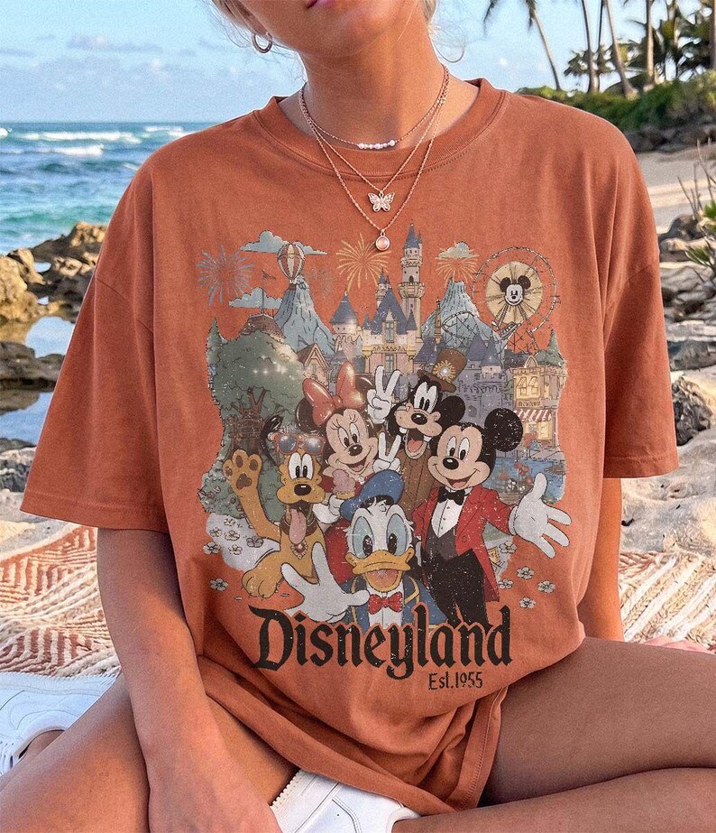 May include: A rust-colored short-sleeve t-shirt featuring a graphic print of Mickey Mouse, Minnie Mouse, Donald Duck, Goofy, and Pluto in front of a castle. The text "Disneyland Est. 1955" is printed in black at the bottom of the graphic design.