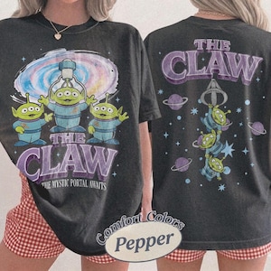 May include: A charcoal gray t-shirt featuring a graphic of three green three-eyed aliens under a claw machine. The front displays the text THE CLAW THE MYSTIC PORTAL AWAITS. The back shows the aliens hanging from a claw with the text THE CLAW.