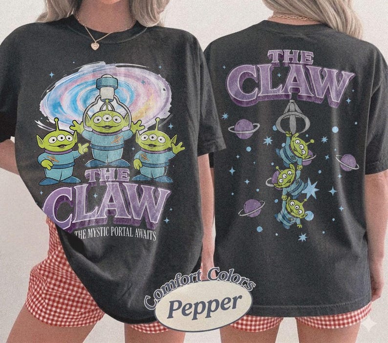 May include: A charcoal gray t-shirt featuring a graphic of three green three-eyed aliens under a claw machine. The front displays the text THE CLAW THE MYSTIC PORTAL AWAITS. The back shows the aliens hanging from a claw with the text THE CLAW.