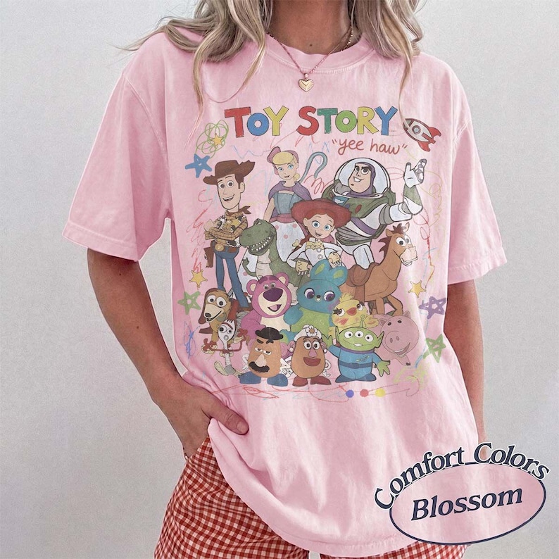 May include: A light pink short-sleeve t-shirt featuring a colorful graphic print of Toy Story characters including Woody, Buzz Lightyear, Jessie, and others. The text "TOY STORY" and "yee haw" are printed above the characters with small star doodles.