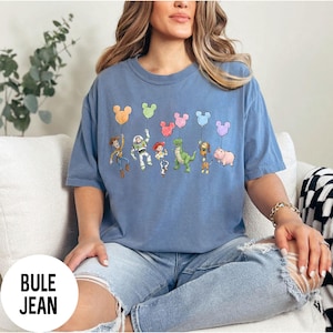 K&ouml;nnte beinhalten: Hellblaues T-Shirt mit einem Cartoon-Motiv von Zeichentrickfiguren, die Mickey Mouse-f&ouml;rmige Ballons halten. Der Text "BULE JEAN" befindet sich in einem wei&szlig;en Kreis unten links. Das T-Shirt hat eine lockere Passform.