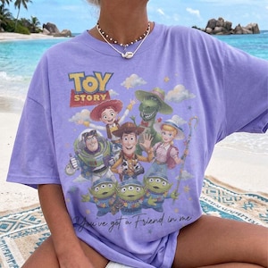 Puede incluir: Una camiseta de color morado claro con un estampado gr&aacute;fico de los personajes de Toy Story, incluyendo a Woody, Buzz Lightyear, Jessie, Bo Peep, Rex y los Aliens. El texto Toy Story aparece arriba y la frase You've got a Friend in me debajo.