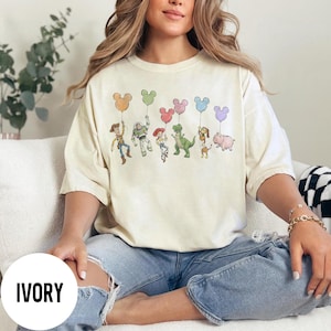 K&ouml;nnte beinhalten: Elfenbeinfarbenes T-Shirt mit einer Grafik von Zeichentrickfiguren, die Mickey Mouse-f&ouml;rmige Ballons halten. Zu den Figuren geh&ouml;ren Woody, Buzz Lightyear, Jessie, Rex, Slinky Dog und Hamm. Das Wort "IVORY" ist auf einem wei&szlig;en Kreis unten aufgedruckt.
