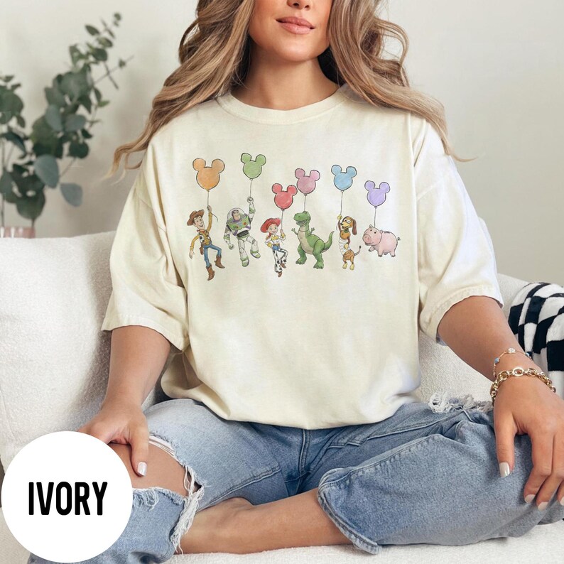 K&ouml;nnte beinhalten: Elfenbeinfarbenes T-Shirt mit einer Grafik von Zeichentrickfiguren, die Mickey Mouse-f&ouml;rmige Ballons halten. Zu den Figuren geh&ouml;ren Woody, Buzz Lightyear, Jessie, Rex, Slinky Dog und Hamm. Das Wort "IVORY" ist auf einem wei&szlig;en Kreis unten aufgedruckt.