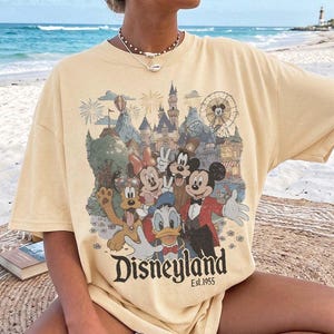 May include: A pale yellow short-sleeve t-shirt featuring a graphic print of Mickey Mouse, Minnie Mouse, Donald Duck, Goofy, and Pluto in front of a castle and ferris wheel. The text Disneyland Est. 1955 is printed in black at the bottom of the graphic.