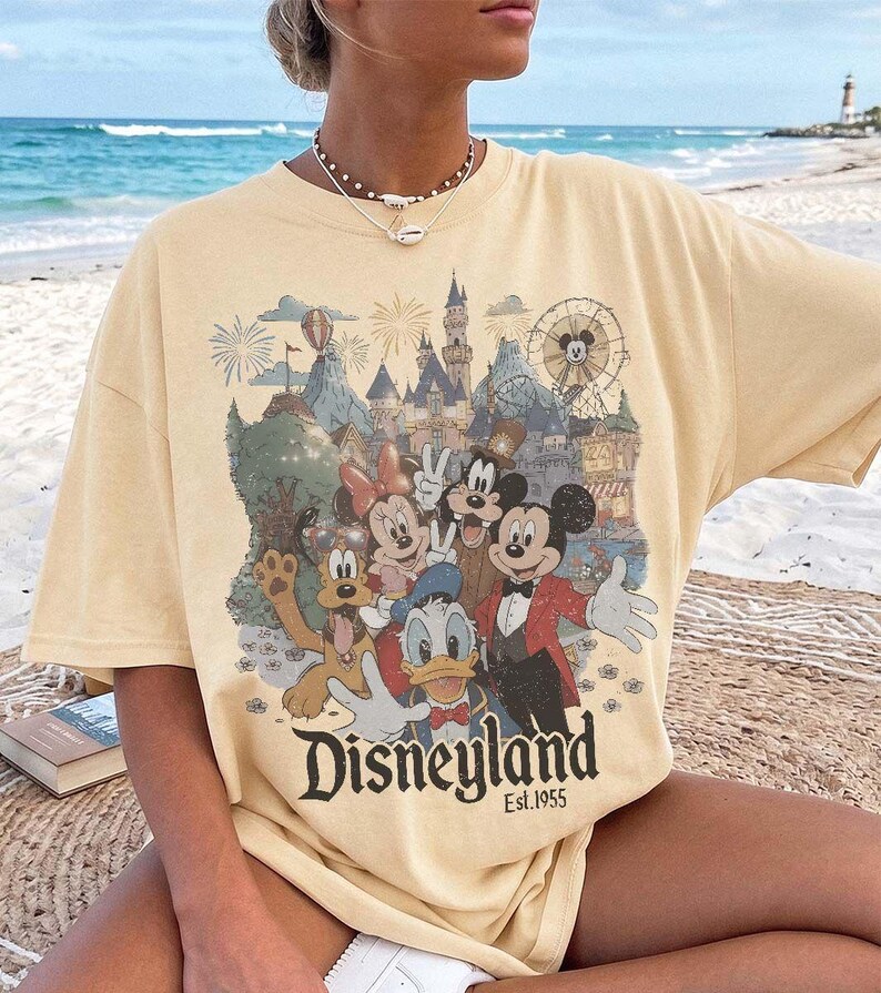May include: A pale yellow short-sleeve t-shirt featuring a graphic print of Mickey Mouse, Minnie Mouse, Donald Duck, Goofy, and Pluto in front of a castle and ferris wheel. The text Disneyland Est. 1955 is printed in black at the bottom of the graphic.