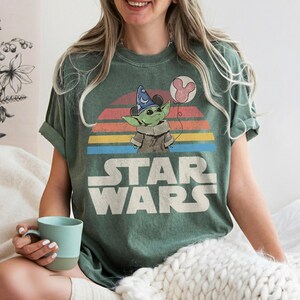 May include: Green t-shirt featuring a graphic of Grogu from Star Wars wearing a wizard hat and holding a Mickey Mouse balloon. The Star Wars logo is printed below the graphic, with a colorful sunset design in the background.