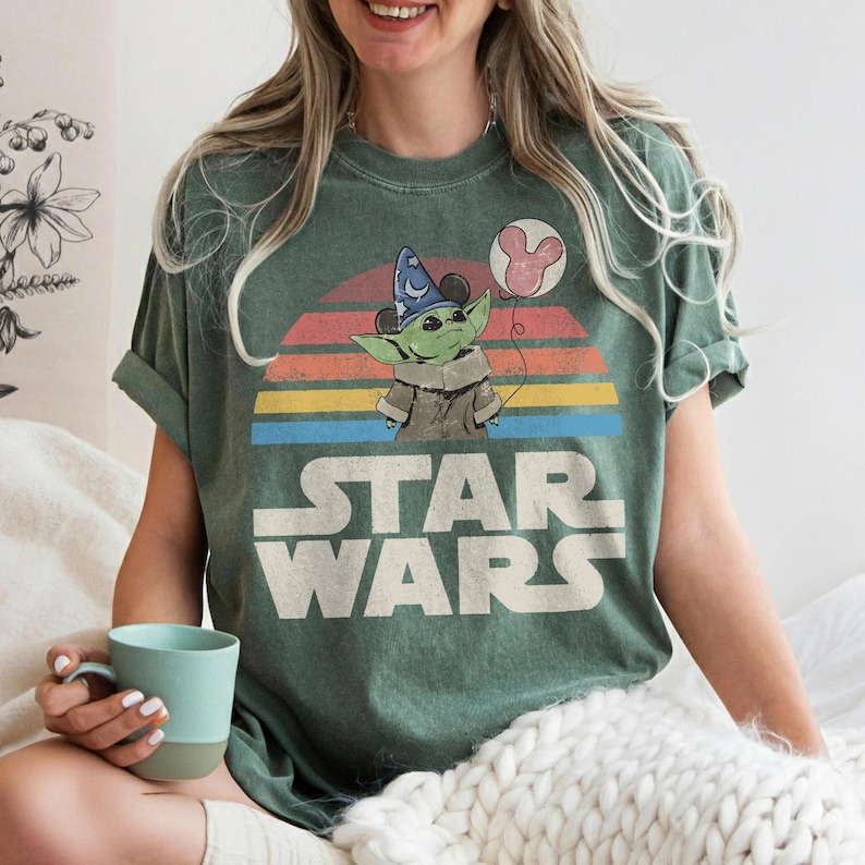 May include: Green t-shirt featuring a graphic of Grogu from Star Wars wearing a wizard hat and holding a Mickey Mouse balloon. The Star Wars logo is printed below the graphic, with a colorful sunset design in the background.