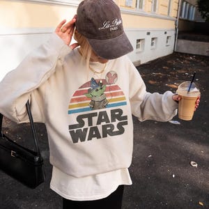 May include: Cream-colored Star Wars sweatshirt featuring a graphic of Grogu with a wizard hat and a Mickey Mouse balloon against a sunset design. The words "STAR WARS" are printed in large, bold letters below the graphic.