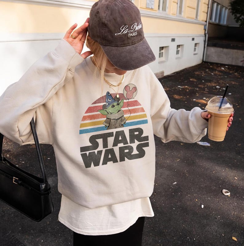 May include: Cream-colored Star Wars sweatshirt featuring a graphic of Grogu with a wizard hat and a Mickey Mouse balloon against a sunset design. The words "STAR WARS" are printed in large, bold letters below the graphic.