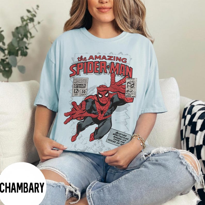 May include: Light blue t-shirt featuring a comic book cover design of Spider-Man. The design includes the text "the AMAZING SPIDER-MAN" and "MARVEL COMICS GROUP". The superhero is depicted in a dynamic pose with outstretched hands.