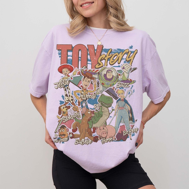 May include: A light purple short-sleeve t-shirt featuring a large graphic print of Toy Story characters including Woody, Buzz Lightyear, Jessie, Rex, Slinky Dog, Bullseye, Hamm, and Bo Peep, with the text Toy Story printed in large red and yellow letters.