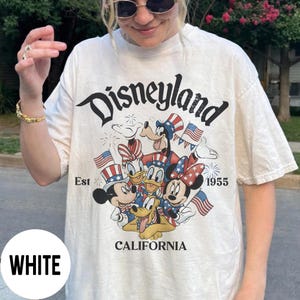 May include: White t-shirt featuring the word "Disneyland" in a black script font. The shirt has a graphic of Mickey Mouse, Minnie Mouse, Donald Duck, Daisy Duck, and Goofy in patriotic attire. The text "Est 1955" and "California" are also printed on the shirt.