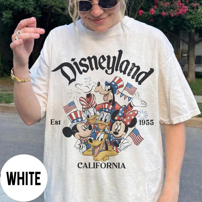 May include: White t-shirt featuring the word "Disneyland" in a black script font. The shirt has a graphic of Mickey Mouse, Minnie Mouse, Donald Duck, Daisy Duck, and Goofy in patriotic attire. The text "Est 1955" and "California" are also printed on the shirt.