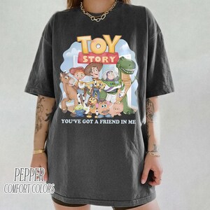 K&ouml;nnte beinhalten: Dunkelgraues T-Shirt mit dem Toy Story-Logo und -Charakteren. Die Grafik zeigt Woody, Buzz Lightyear und Rex mit dem Text "YOU'VE GOT A FRIEND IN ME". Das Shirt ist von Pepper Comfort Colors.