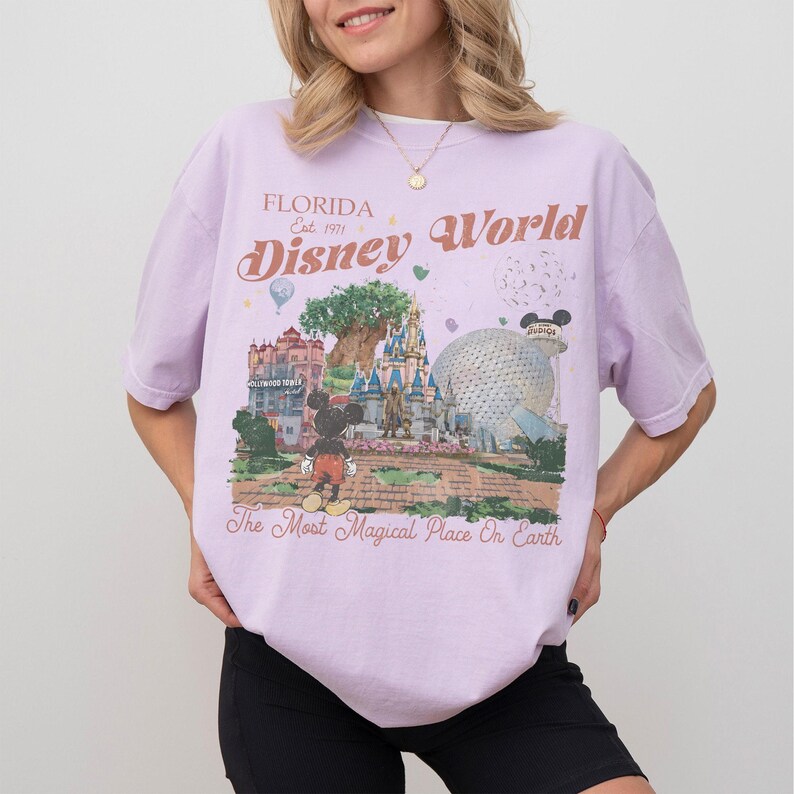 May include: A light purple t-shirt with a Disney World graphic. The graphic includes the text "Florida Est. 1971 Disney World" and an illustration of Mickey Mouse, a castle, and Spaceship Earth. The shirt also has the text "The Most Magical Place On Earth."