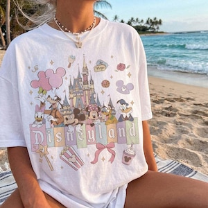May include: A white short-sleeve t-shirt featuring a colourful graphic print of a castle, various cartoon characters, and the text Disneyland. The design includes pastel-coloured accents, balloons, a bow, popcorn, and a drink cup illustration.