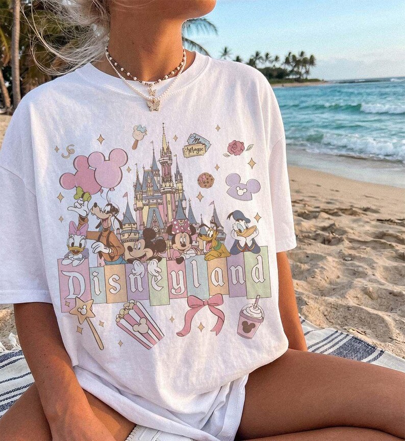 May include: A white short-sleeve t-shirt featuring a colourful graphic print of a castle, various cartoon characters, and the text Disneyland. The design includes pastel-coloured accents, balloons, a bow, popcorn, and a drink cup illustration.