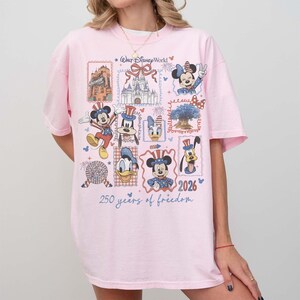 Może przedstawiać: Jasnor&oacute;żowy t-shirt z nadrukiem graficznym postaci z Walt Disney World, w tym Myszki Miki, Myszki Minnie, Kaczora Donalda, Kaczki Daisy i Goofy'ego. Projekt zawiera tekst Walt Disney World, 2026 oraz 250 years of freedom.