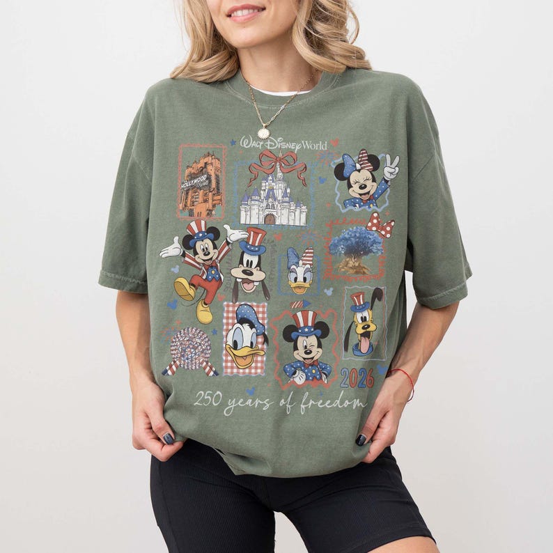 Może przedstawiać: Oliwkowozielony t-shirt z nadrukiem graficznym zawierającym branding Walt Disney World, tekst 250 years of freedom oraz 2026. Wz&oacute;r zawiera ilustracje Myszki Miki, Myszki Minnie, Kaczora Donalda, Goofy'ego i Pluto w patriotycznych strojach.