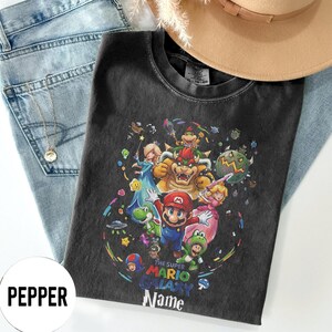 Puede incluir: Camiseta negra con un gr&aacute;fico colorido de personajes del videojuego Super Mario Galaxy, incluyendo a Mario, Yoshi y Bowser. El dise&ntilde;o est&aacute; rodeado de estrellas y planetas. El texto "The Super Mario Galaxy Name" tambi&eacute;n es visible.
