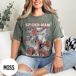 Pu&ograve; includere: T-shirt verde muschio con grafica vintage di Spider-Man. Il design include Spider-Man che si dondola, la scritta "Spider-Man New York" e l'anno "1962". La maglietta ha un'estetica retr&ograve;.