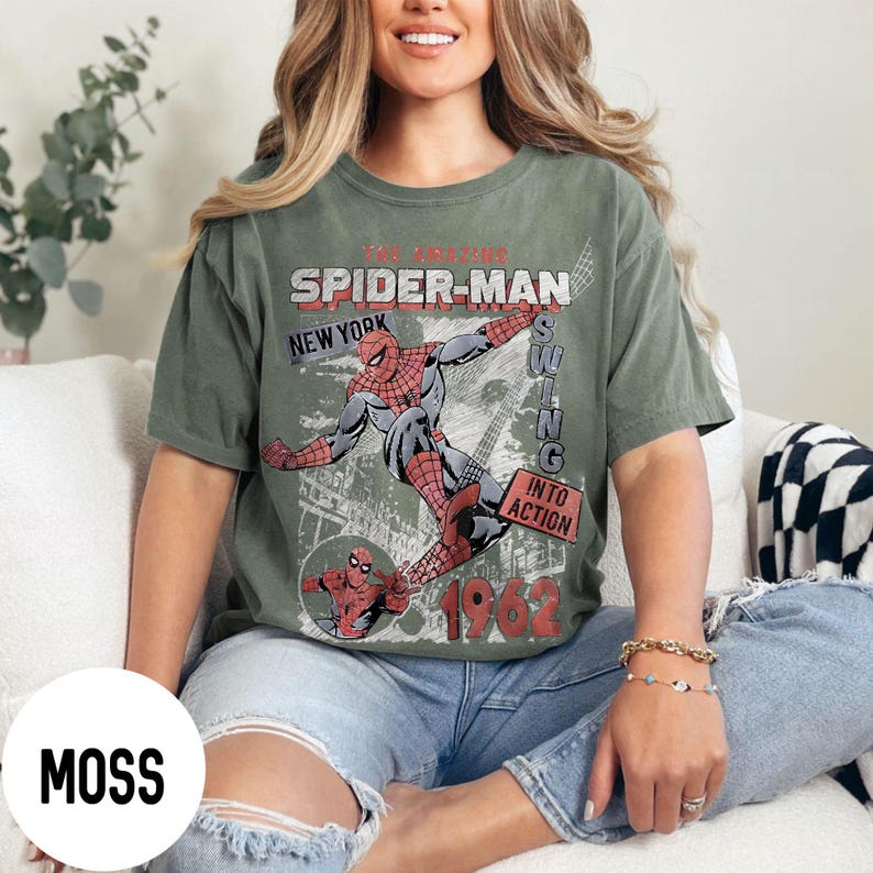 Pu&ograve; includere: T-shirt verde muschio con grafica vintage di Spider-Man. Il design include Spider-Man che si dondola, la scritta "Spider-Man New York" e l'anno "1962". La maglietta ha un'estetica retr&ograve;.