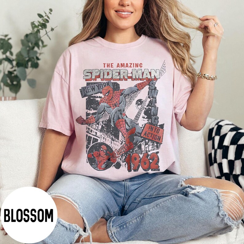 Pu&ograve; includere: T-shirt rosa chiaro con grafica vintage di Spider-Man. Il design include il testo "THE AMAZING SPIDER-MAN", "NEW YORK" e "1962". La grafica mostra Spider-Man in azione, con uno sfondo di paesaggio urbano.