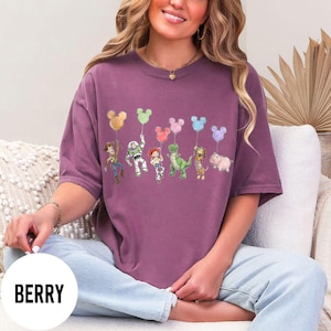 K&ouml;nnte beinhalten: Beerenfarbenes T-Shirt mit einem Cartoon-Design von Figuren, die Mickey Mouse-f&ouml;rmige Ballons halten. Die Figuren sind Woody, Buzz Lightyear, Jessie, Rex, Slinky Dog und Hamm. Das Wort "BERRY" ist auf einem wei&szlig;en Kreis aufgedruckt.