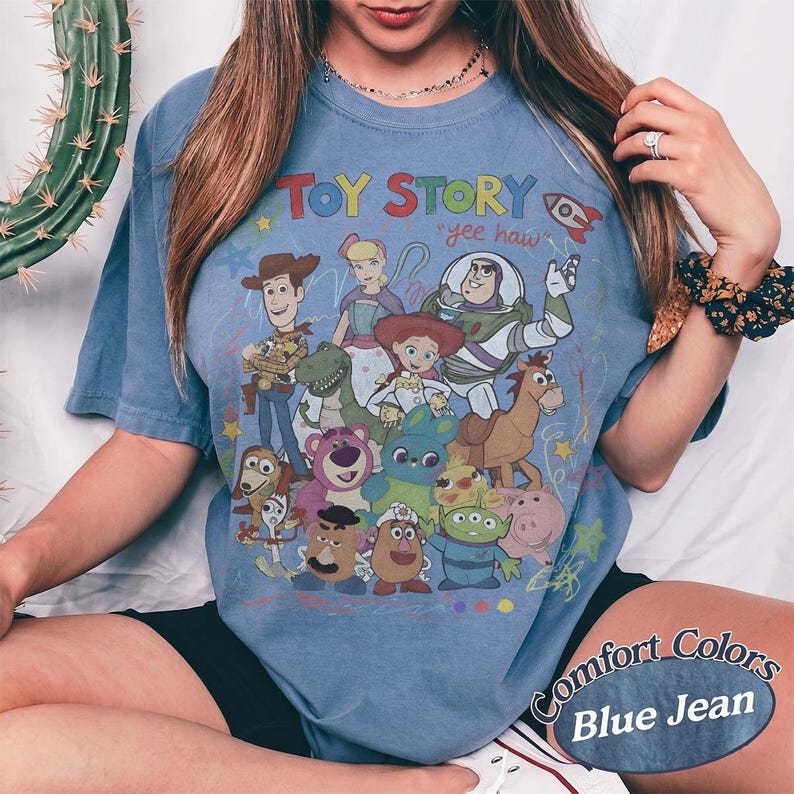 May include: A blue short-sleeve t-shirt featuring a colorful graphic print of Toy Story characters including Woody, Buzz Lightyear, Jessie, and others. The text "TOY STORY" and "yee haw" is printed above the characters in a playful, multi-colored font.