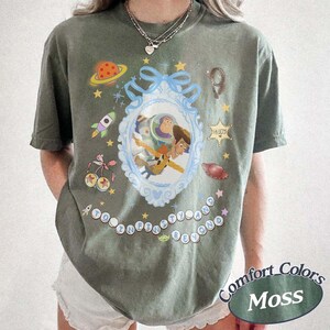 Peut inclure: Un t-shirt &agrave; manches courtes vert mousse avec un cadre orn&eacute; bleu clair central contenant des illustrations de Buzz l'&Eacute;clair et Woody. Autour du cadre se trouvent de petits graphismes d'une plan&egrave;te, d'une fus&eacute;e, d'&eacute;toiles et le texte "To Infinity and Beyond."