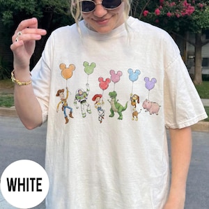 K&ouml;nnte beinhalten: Wei&szlig;es T-Shirt mit einem farbenfrohen Aufdruck von Toy Story-Charakteren, die Mickey Mouse-f&ouml;rmige Ballons halten. Die Charaktere sind Woody, Buzz Lightyear, Jessie, Rex, Slinky Dog und Hamm.