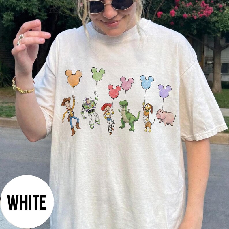 K&ouml;nnte beinhalten: Wei&szlig;es T-Shirt mit einem farbenfrohen Aufdruck von Toy Story-Charakteren, die Mickey Mouse-f&ouml;rmige Ballons halten. Die Charaktere sind Woody, Buzz Lightyear, Jessie, Rex, Slinky Dog und Hamm.