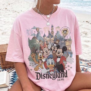 May include: A light pink short-sleeved t-shirt featuring a graphic print of Mickey Mouse, Minnie Mouse, Donald Duck, Goofy, and Pluto in front of a castle and ferris wheel. The text "Disneyland Est. 1955" is printed in black below the characters.