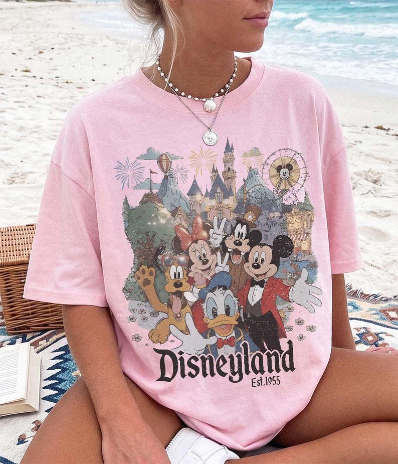 May include: A light pink short-sleeved t-shirt featuring a graphic print of Mickey Mouse, Minnie Mouse, Donald Duck, Goofy, and Pluto in front of a castle and ferris wheel. The text "Disneyland Est. 1955" is printed in black below the characters.