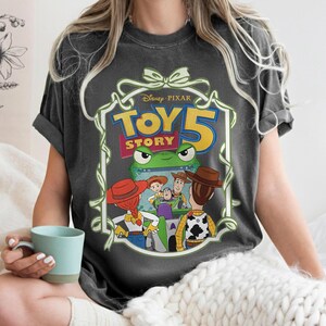 May include: Dark gray t-shirt featuring a colorful graphic from Disney Pixar's Toy Story 5. The design includes the movie title, a green frog, and several characters. The shirt has short sleeves and a crew neck.