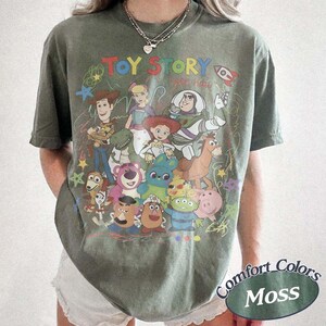 May include: A moss green short-sleeve t-shirt featuring a colorful graphic print of Toy Story characters including Woody, Buzz Lightyear, Jessie, and others. The text TOY STORY is printed in multicolored letters above the character illustration.