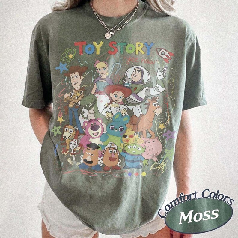 May include: A moss green short-sleeve t-shirt featuring a colorful graphic print of Toy Story characters including Woody, Buzz Lightyear, Jessie, and others. The text TOY STORY is printed in multicolored letters above the character illustration.