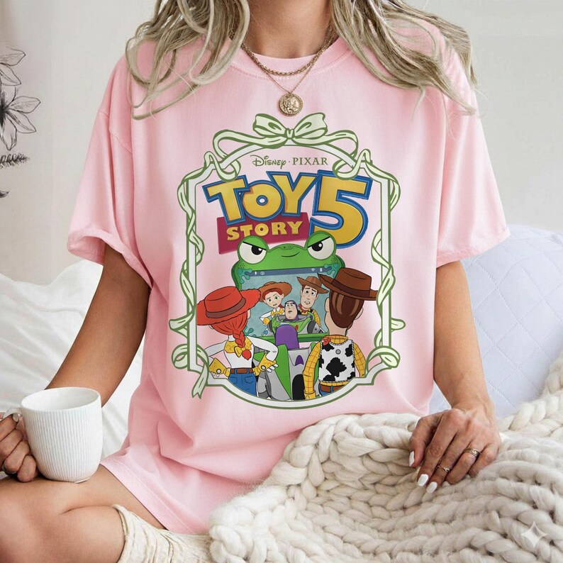 May include: A light pink t-shirt featuring a graphic from the Disney Pixar movie Toy Story 5. The graphic includes the movie title, a green frog, and several characters from the film. The shirt is short-sleeved.