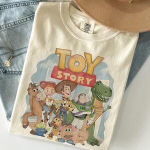 K&ouml;nnte beinhalten: Cremefarbenes T-Shirt mit dem Schriftzug "TOY STORY" in einer farbenfrohen Grafik. Die Grafik zeigt Zeichentrickfiguren wie Woody, Buzz Lightyear und Rex. Das T-Shirt ist auf einer blauen Jeans und einem braunen Hut gefaltet.