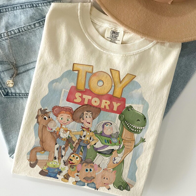 K&ouml;nnte beinhalten: Cremefarbenes T-Shirt mit dem Schriftzug "TOY STORY" in einer farbenfrohen Grafik. Die Grafik zeigt Zeichentrickfiguren wie Woody, Buzz Lightyear und Rex. Das T-Shirt ist auf einer blauen Jeans und einem braunen Hut gefaltet.