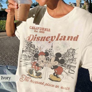 May include: Ivory-colored t-shirt featuring a vintage Disneyland graphic. The design includes Mickey and Minnie Mouse, fireworks, and the text "CALIFORNIA Est. 1955 Disneyland The happiest place on earth."
