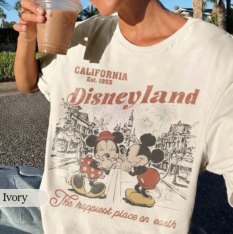 May include: Ivory-colored t-shirt featuring a vintage Disneyland graphic. The design includes Mickey and Minnie Mouse, fireworks, and the text "CALIFORNIA Est. 1955 Disneyland The happiest place on earth."
