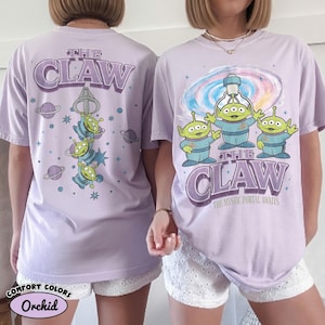 May include: A light purple t-shirt featuring a graphic of three green three-eyed aliens. The front displays the text THE CLAW and THE MYSTIC PORTAL AWAITS with a swirling portal. The back shows the aliens hanging from a claw with planet icons.