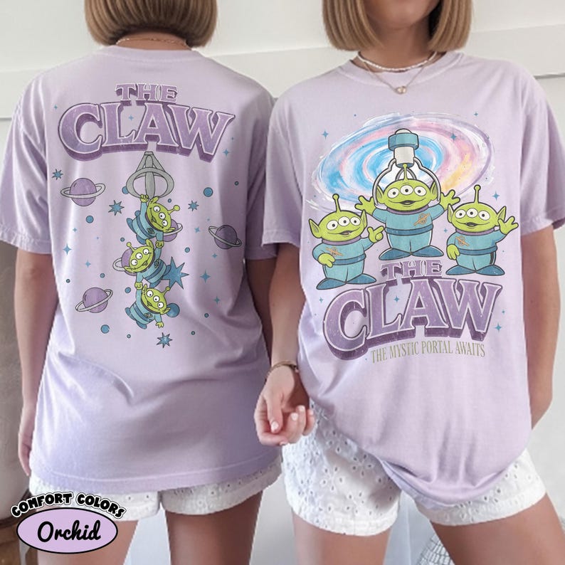 May include: A light purple t-shirt featuring a graphic of three green three-eyed aliens. The front displays the text THE CLAW and THE MYSTIC PORTAL AWAITS with a swirling portal. The back shows the aliens hanging from a claw with planet icons.